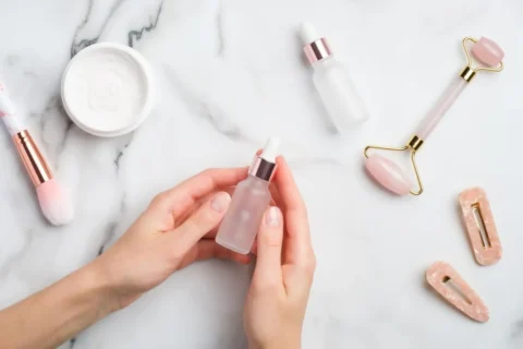 Female hand holding serum bottle over marble table with moisturizer cream makeup brush face massage roller