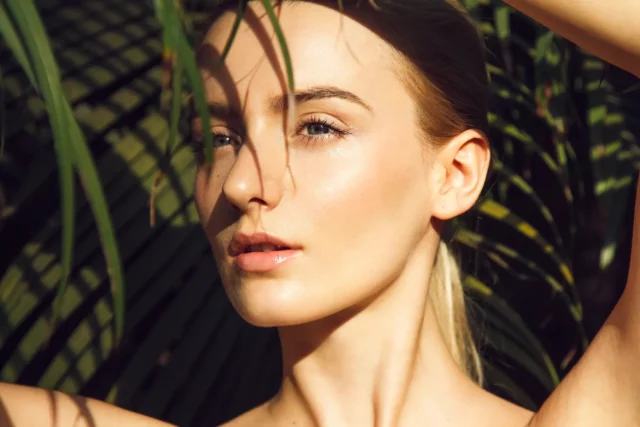 a portrait of a young, blonde woman with perfect skin hiding under a palm leaf
